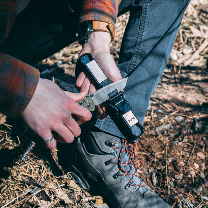 A close-up outdoor shot of a person sharpening a knife using a handheld Venture knife sharpener over a hiking boot on a forest floor covered in dry leaves.