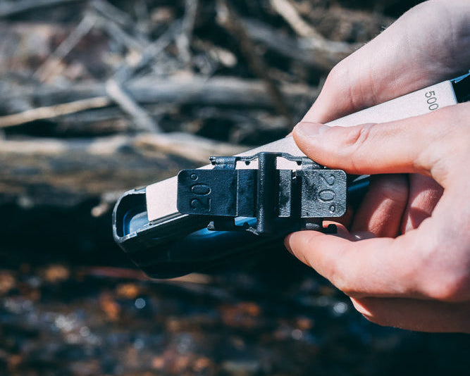 A close-up of a person's hands holding 20° knife guide. They are attaching it to the Venture knife sharpener set against a blurred natural background of logs and water.