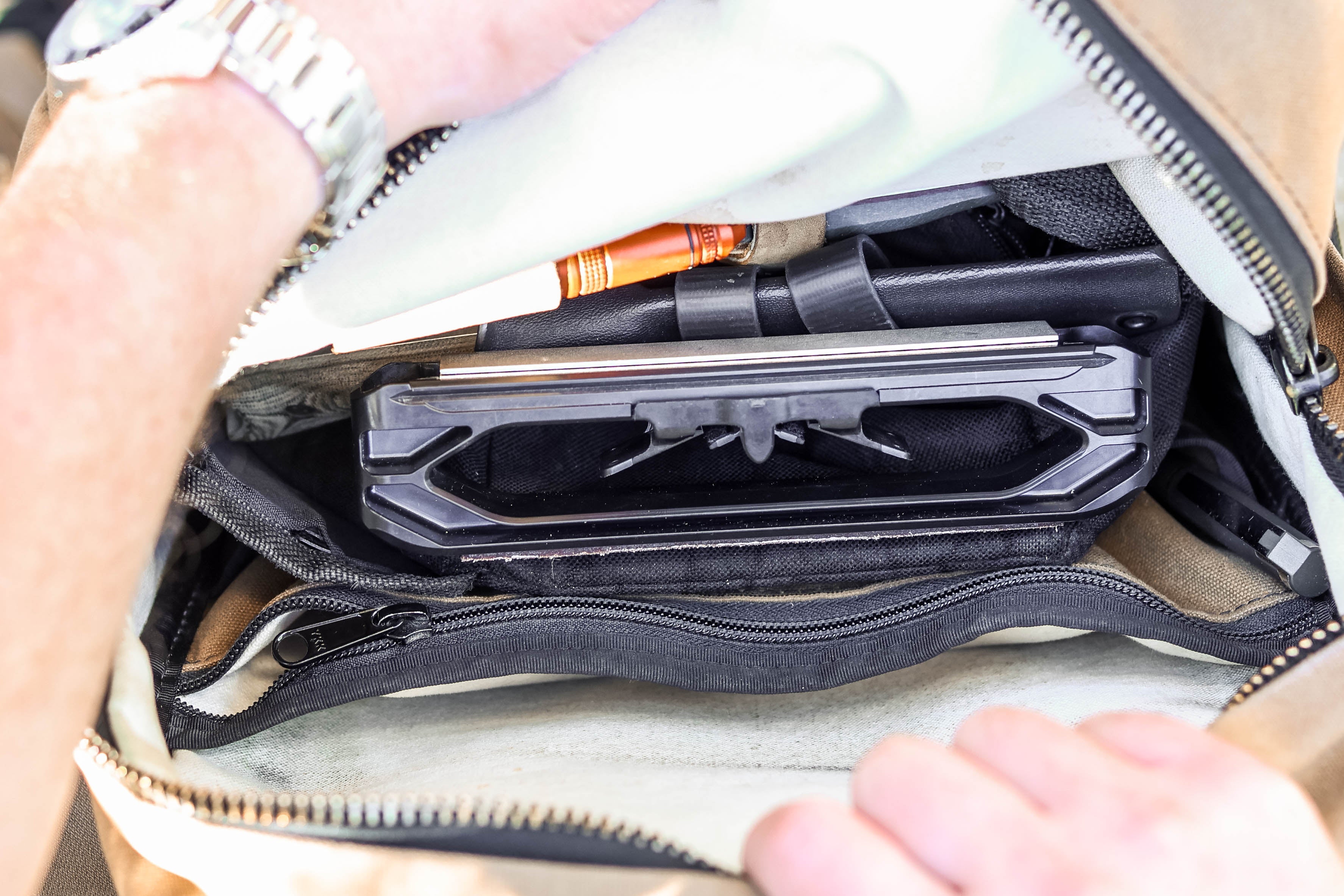 Close-up of a tan backpack or duffel bag being opened by two hands, showing a black padded laptop sleeve and other gear inside.