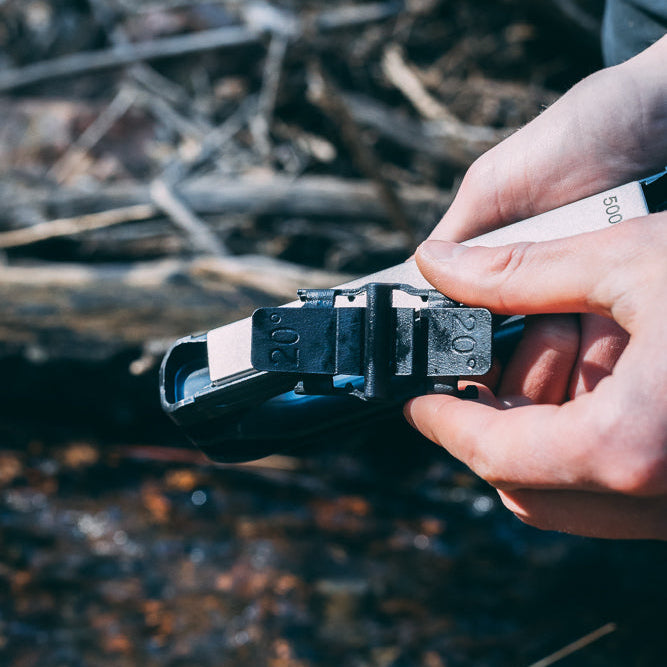 A close-up of a person's hands holding 20° knife guide. They are attaching it to the Venture knife sharpener set against a blurred natural background of logs and water.