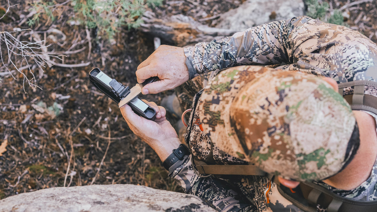 A person in camouflage hunting clothing kneels on rocky, wooded ground while holding a Venture knife sharpener sharpening a knife