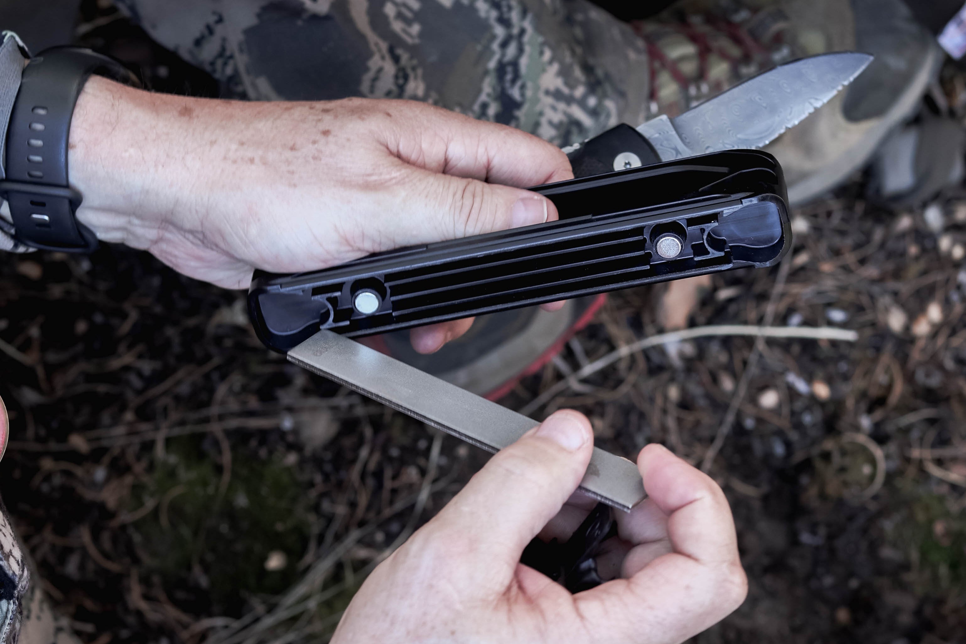 A close-up of a person outdoors holding a black multi-tool pocket knife with silver blades partially extended, wearing camouflage clothing and a black watch.